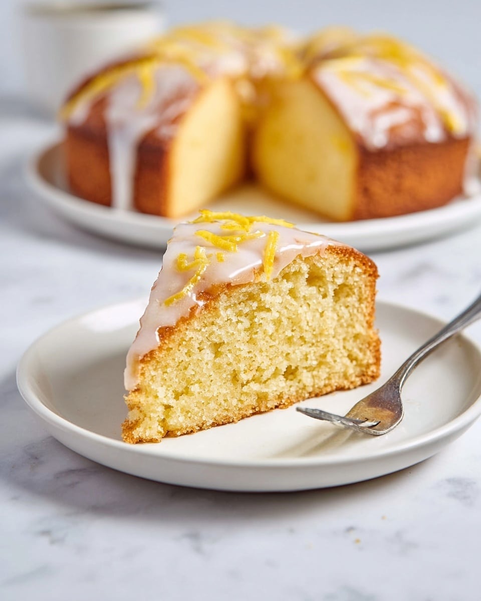 The image shows one slice of lemon cake placed on a white plate with a silver fork beside it. The cake has a golden-brown crust on the sides and a light yellow, moist texture inside. On top, there is a thin layer of white glaze, drizzled unevenly, with thin strips of yellow lemon zest scattered on the glaze. In the background, there is another larger piece of the same lemon cake on a similar white plate, slightly blurred. The setting is on a white marbled surface. photo taken with an iphone --ar 4:5 --v 7