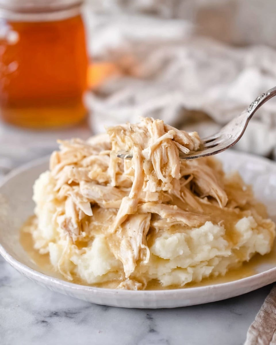 A white round plate on a white marbled surface holds a base layer of creamy mashed potatoes, smooth and slightly fluffy with a soft off-white color. Piled high on top is shredded light brown chicken mixed with thick beige gravy that coats the meat, giving it a glossy texture. A fork lifting a portion of the chicken and gravy reveals tender, stringy pieces dripping slightly, contrasting with the mashed potato base below. In the background, blurred, is a white cloth and a glass jar with an amber-colored liquid, all softly lit. photo taken with an iphone --ar 4:5 --v 7