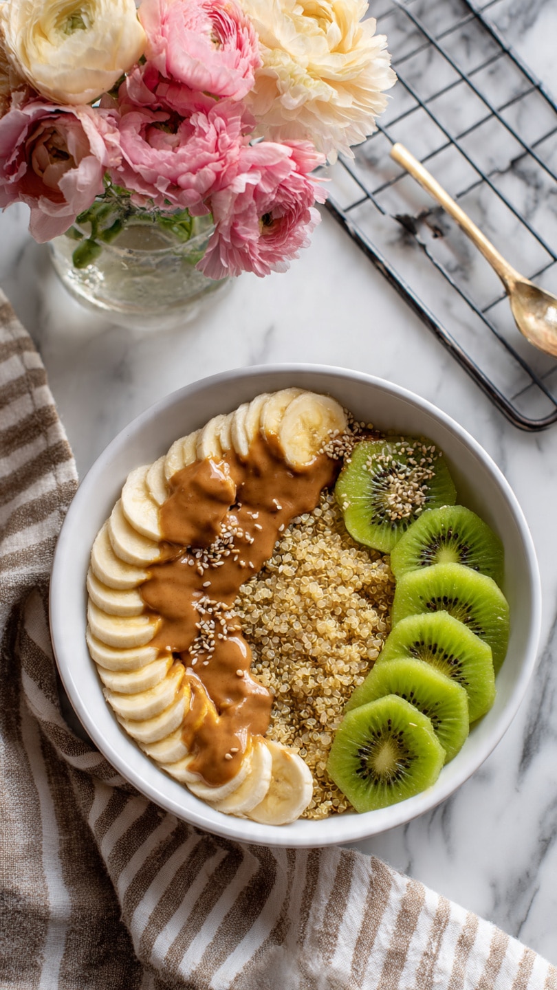 A white bowl filled with a base layer of cooked quinoa, light brown and fluffy in texture. On top of the quinoa, there is a neat row of thin banana slices, pale yellow and smooth, positioned along the edge of the bowl. In the center, several pieces of bright green kiwi slices add a fresh contrast. Drizzled over the fruit and quinoa is a light brown nut butter, slightly shiny and creamy. Small white and black seeds are sprinkled over everything, adding texture. The bowl is on a white marbled surface with a metal cooling rack and a beige cloth with stripes in the background, along with a glass vase holding pink and cream flowers. Photo taken with an iphone --ar 4:5 --v 7