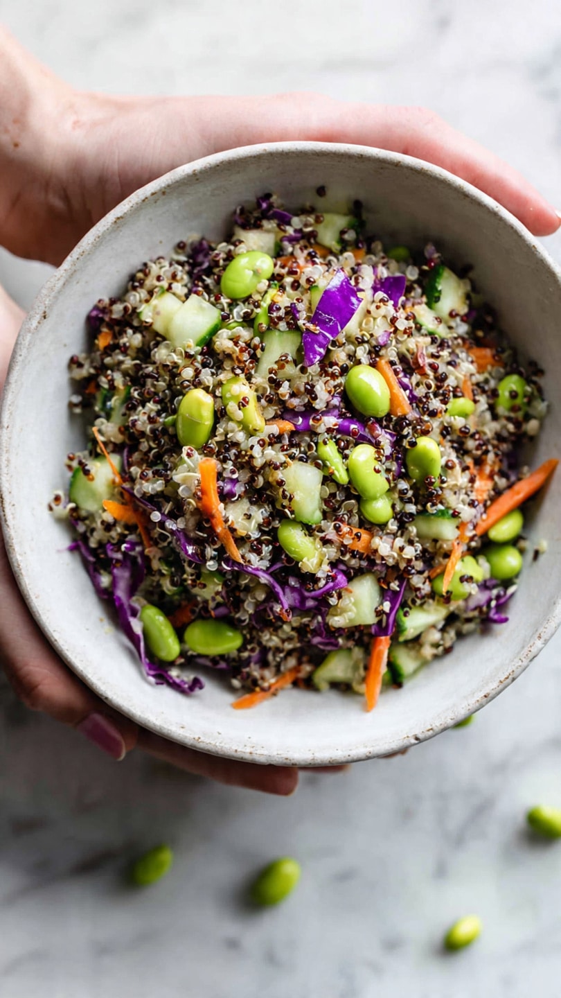 A close-up view of a white bowl filled with a colorful quinoa salad. The salad layers show a mix of small, fluffy quinoa grains in white, black, and beige colors as the base. On top, there are bright green edamame beans, diced green cucumber pieces, thin orange carrot strips, and bits of purple cabbage for contrast. The bowl sits on a white marbled surface with some scattered edamame beans around it. A woman's hand is gently holding the side of the bowl. Photo taken with an iphone --ar 4:5 --v 7