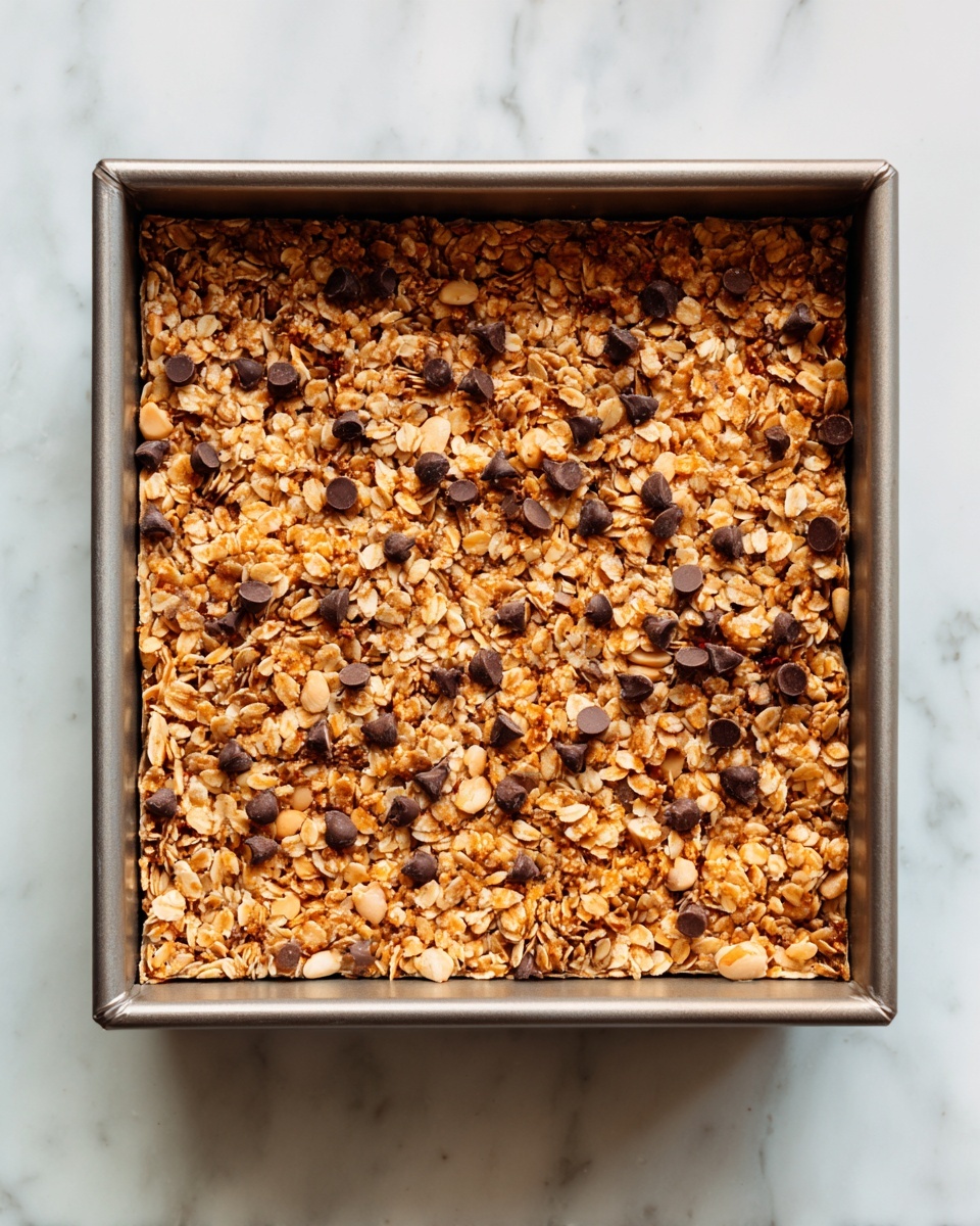 A square baking pan filled with a single layer of homemade granola bars. The granola has a mix of light golden oats and peanuts, creating a rough texture. Small dark chocolate chips are scattered evenly across the top, providing a contrast against the lighter granola base. The pan edges and corners are visible, showing the granola filling it fully but not overflowing. It sits on a white marbled surface. Photo taken with an iphone --ar 4:5 --v 7