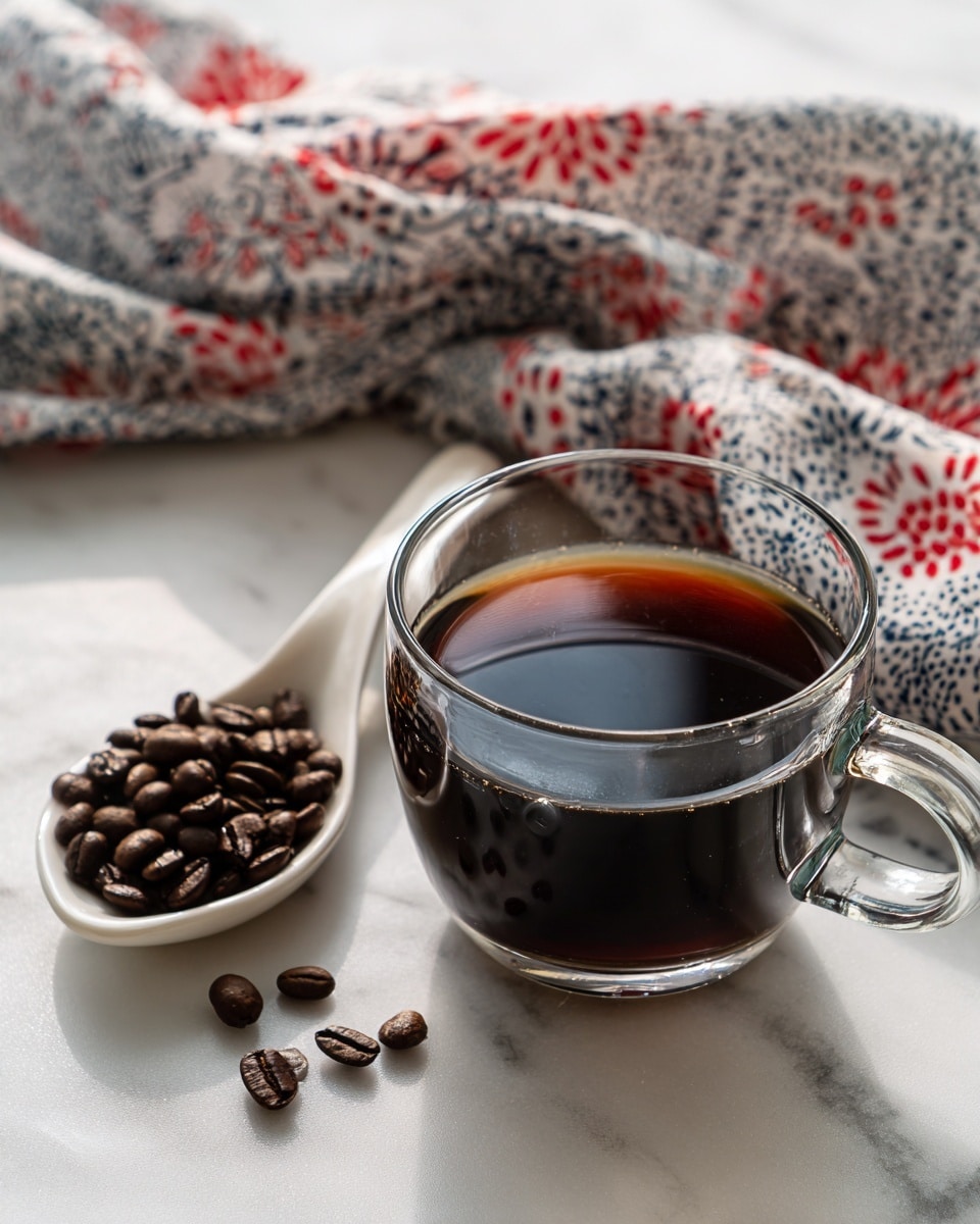 A clear glass cup filled with dark brown coffee sits on a white marbled surface. To the left of the cup, there is a white ceramic spoon holding a small pile of dark brown coffee beans. Behind the cup and spoon, a cloth with a red, white, and dark blue pattern is casually placed, adding a soft texture and color contrast to the scene. The glass cup has a visible handle on the right side. Photo taken with an iphone --ar 4:5 --v 7