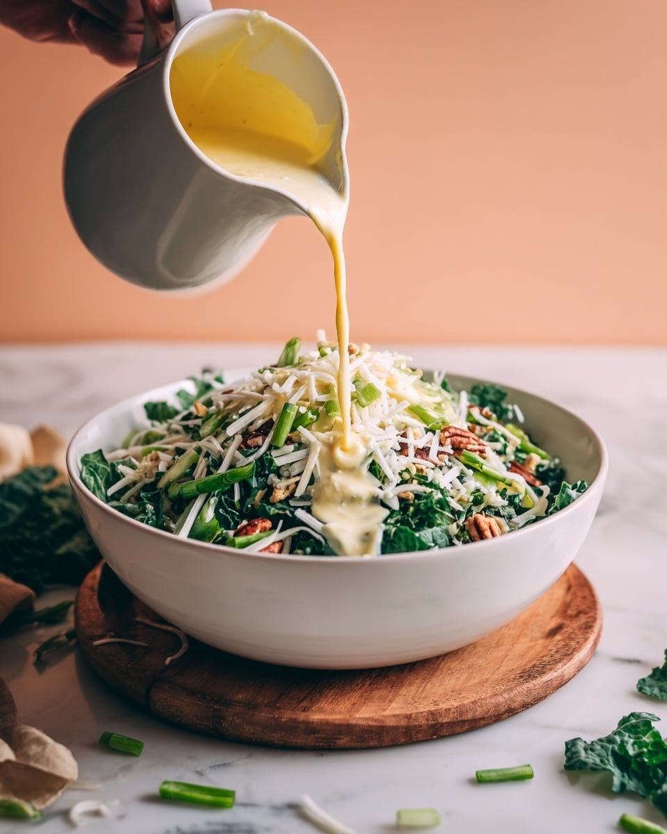 A large white bowl filled with layers of chopped leafy green vegetables and nuts, topped with white shredded cheese and finely sliced green onions. A yellow creamy sauce is being poured from a white jug over the center mound of the salad, creating a smooth texture on top. The bowl sits on a wooden board with some loose greens on the side, against a soft peach background and a white marbled surface below. A woman's hand holds the white jug while pouring the sauce. photo taken with an iphone --ar 4:5 --v 7