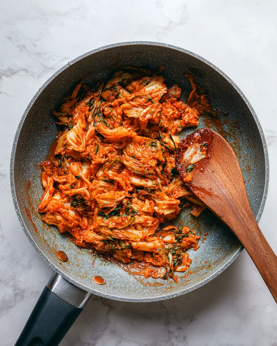 A grey frying pan with a black handle holds a mix of chopped kimchi that is bright red-orange with white and green pieces, showing its spicy and fermented texture. The kimchi is unevenly spread, revealing some shiny, saucy bits and the rough surface of the pan underneath. A wooden spoon lies inside the pan on the right side, partly covered with the kimchi sauce. The background is a white marbled texture. photo taken with an iphone --ar 4:5 --v 7