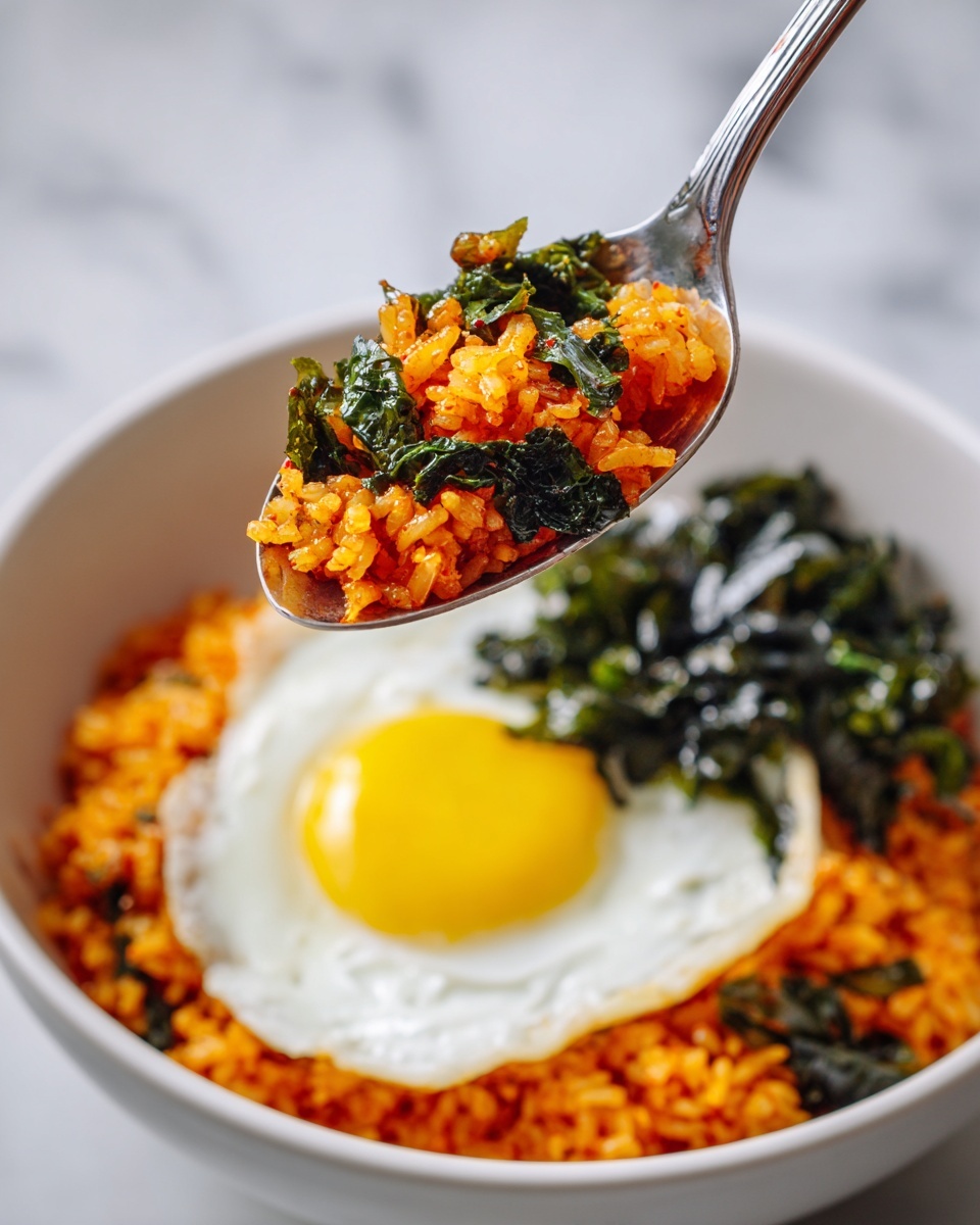 A close-up view of a spoon holding a scoop of spicy orange-red rice mixed with pieces of dark green leafy vegetables and light yellow pickled radish, above a white bowl filled with the same orange rice. To the right inside the bowl is a sunny-side-up egg with a bright yellow yolk and white edges, next to a pile of chopped dark green seaweed. In the background, the bowl sits on a white marbled surface. Photo taken with an iphone --ar 4:5 --v 7