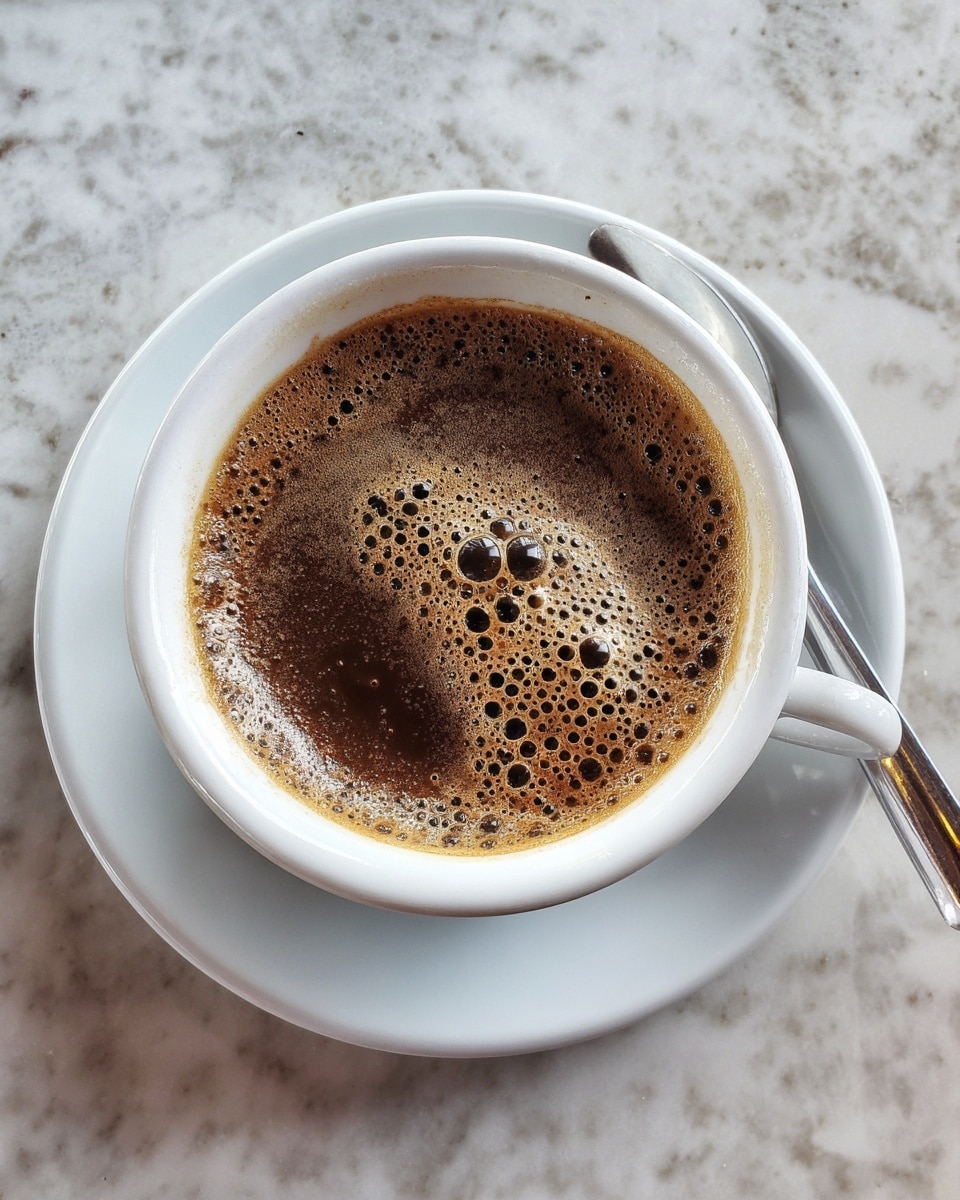 A white cup filled with dark brown coffee topped with a layer of frothy bubbles in shades of deep brown and light tan. The cup sits on a white saucer, all placed on a white marbled surface. Photo taken with an iphone --ar 4:5 --v 7