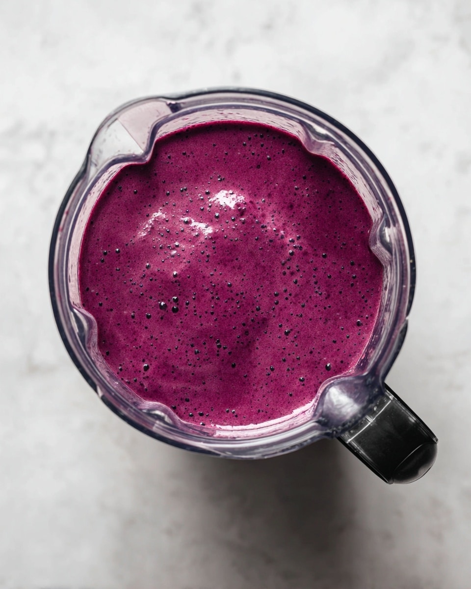 A clear blender jar filled with one smooth layer of thick, bright purple smoothie mixture, slightly shiny with a few tiny bubbles and specks, resting on a white marbled surface. The blender jar has a black handle on the right side. The image is taken from above, showing the full round top opening of the blender jar and its textured contents. photo taken with an iphone --ar 4:5 --v 7