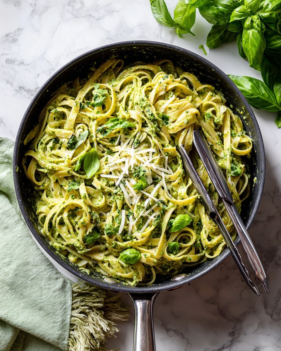 A black pan filled with creamy green pesto sauce coated over tangled fettuccine noodles, with thin pale yellow cheese shavings scattered on top and small fresh green basil pieces sprinkled evenly; silver tongs rest inside the pan holding some pasta. The pan sits on a white marbled surface beside a light green fringed cloth and a bunch of fresh basil leaves. Photo taken with an iphone --ar 4:5 --v 7
