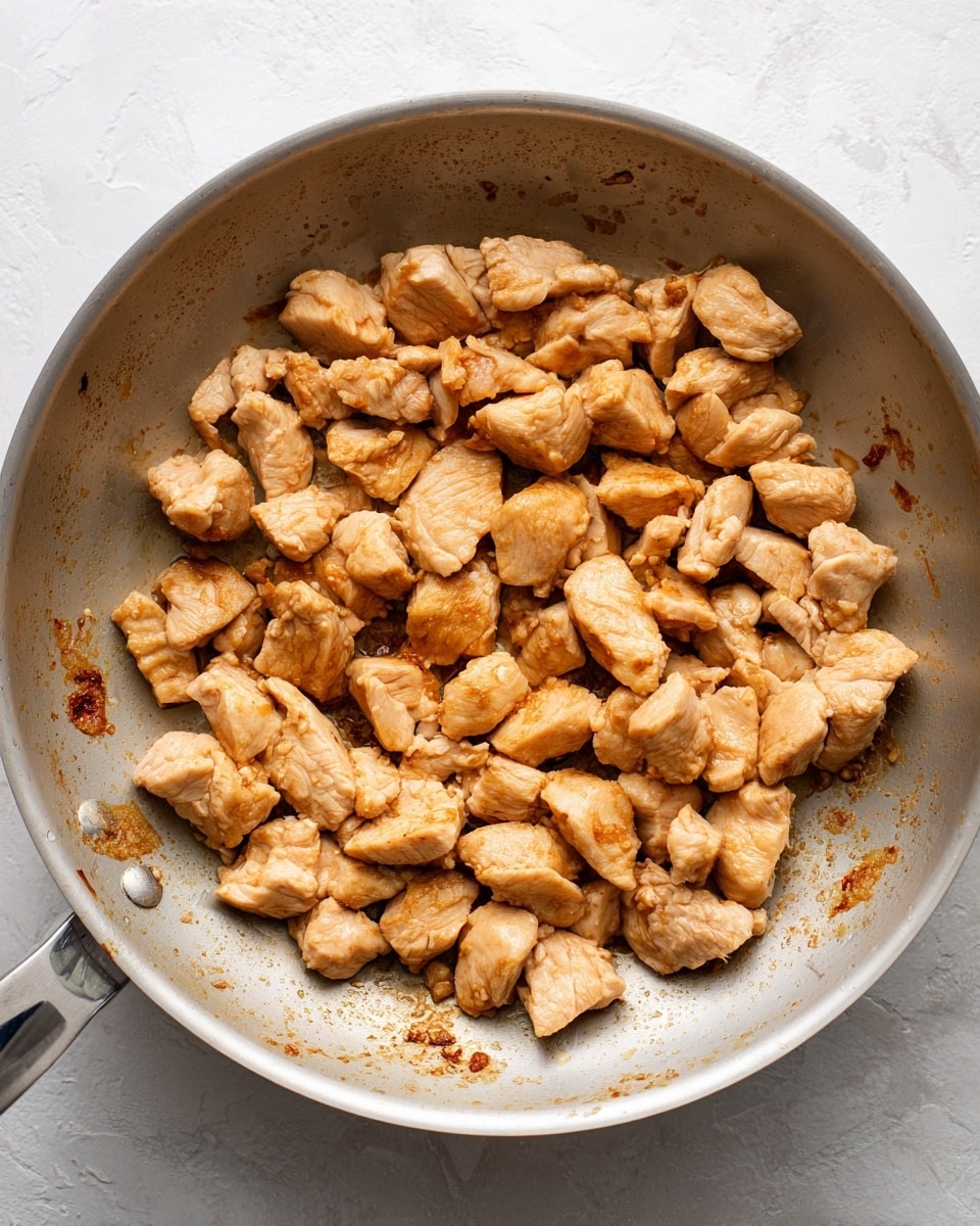 A white metal pan filled with small, cooked pieces of light brown chicken scattered unevenly inside. The chicken pieces have a slightly browned and textured outer surface, with some parts showing a light golden color. The pan shows some browned bits stuck to the surface around the chicken pieces, suggesting cooking residue. The background is a white marbled texture. Photo taken with an iphone --ar 4:5 --v 7