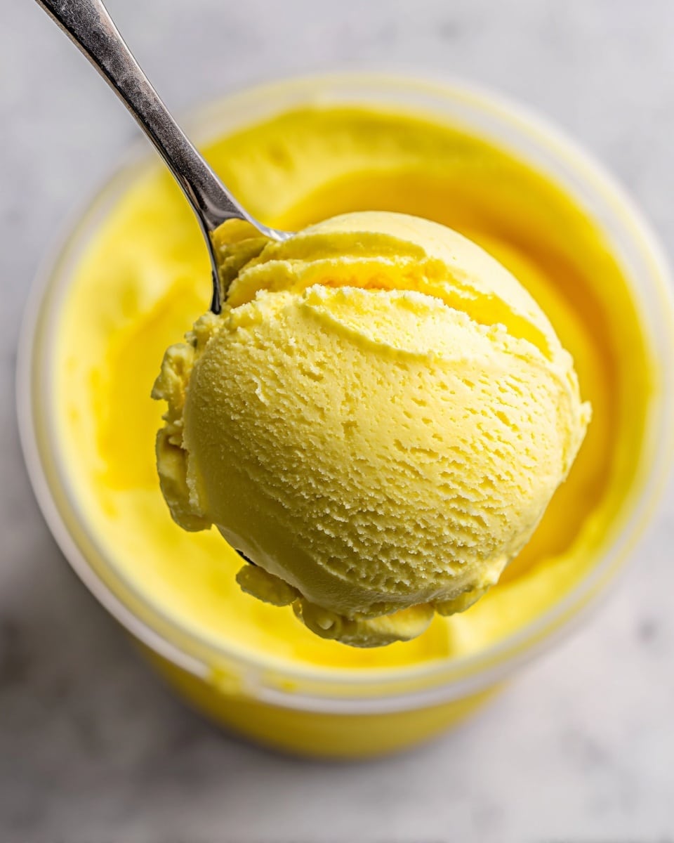 A close-up view of a spoon holding a smooth, creamy scoop of bright yellow ice cream with a slightly soft texture on top, positioned above a clear round container filled with the same yellow ice cream visible inside. The background is a white marbled surface, softly blurred to highlight the ice cream and the spoon in focus. Photo taken with an iphone --ar 4:5 --v 7