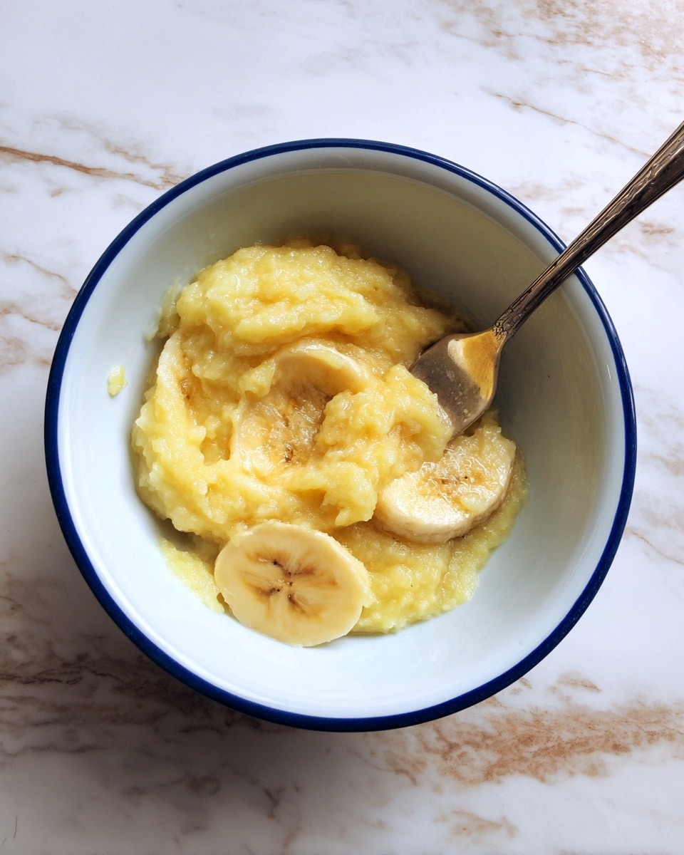 A white bowl with a blue rim holds a small amount of mashed yellowish banana with soft, lumpy texture in the center. A silver fork with a smooth handle rests inside the bowl, partially covered by the mashed banana. The bowl sits on a white marbled surface with faint brown streaks and wood patterns visible around it. Photo taken with an iphone --ar 4:5 --v 7