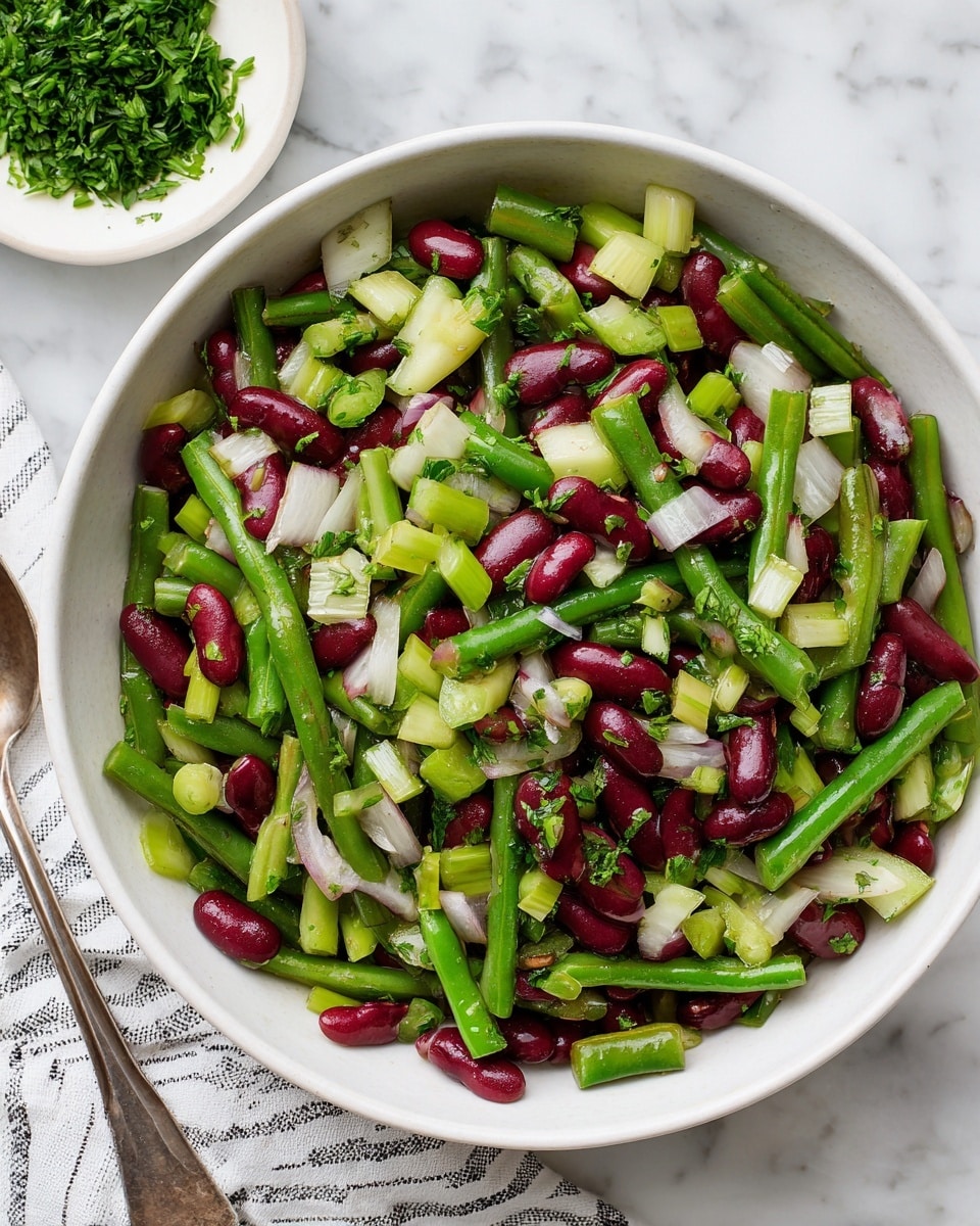 A large white bowl filled with a colorful salad made of chopped green beans, red kidney beans, thin white onion slices, light green celery pieces, and finely chopped green herbs sprinkled on top. The beans are cut into small segments showing a mix of light green and dark green colors. The salad has a shiny, slightly wet texture suggesting a light dressing. The bowl sits on a white marbled surface next to a silver serving spoon in the bottom left corner and a white dish with chopped green herbs in the top area of the image. A white and black striped cloth is partially visible in the lower left corner. Photo taken with an iphone --ar 4:5 --v 7