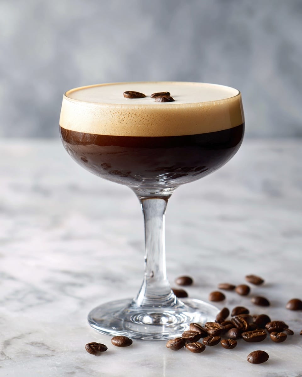 A clear glass coupe holds a two-layer drink with a dark brown bottom layer that looks smooth and rich, topped by a creamy light tan foam layer that fills the glass to the rim. Three whole coffee beans float gently on the foam near the center, adding texture and detail. Around the base of the clear stemmed glass, many scattered coffee beans create a rustic feel. The drink sits on a white marbled surface, enhancing the elegant shape of the glass and the contrasting colors of the drink. photo taken with an iphone --ar 4:5 --v 7