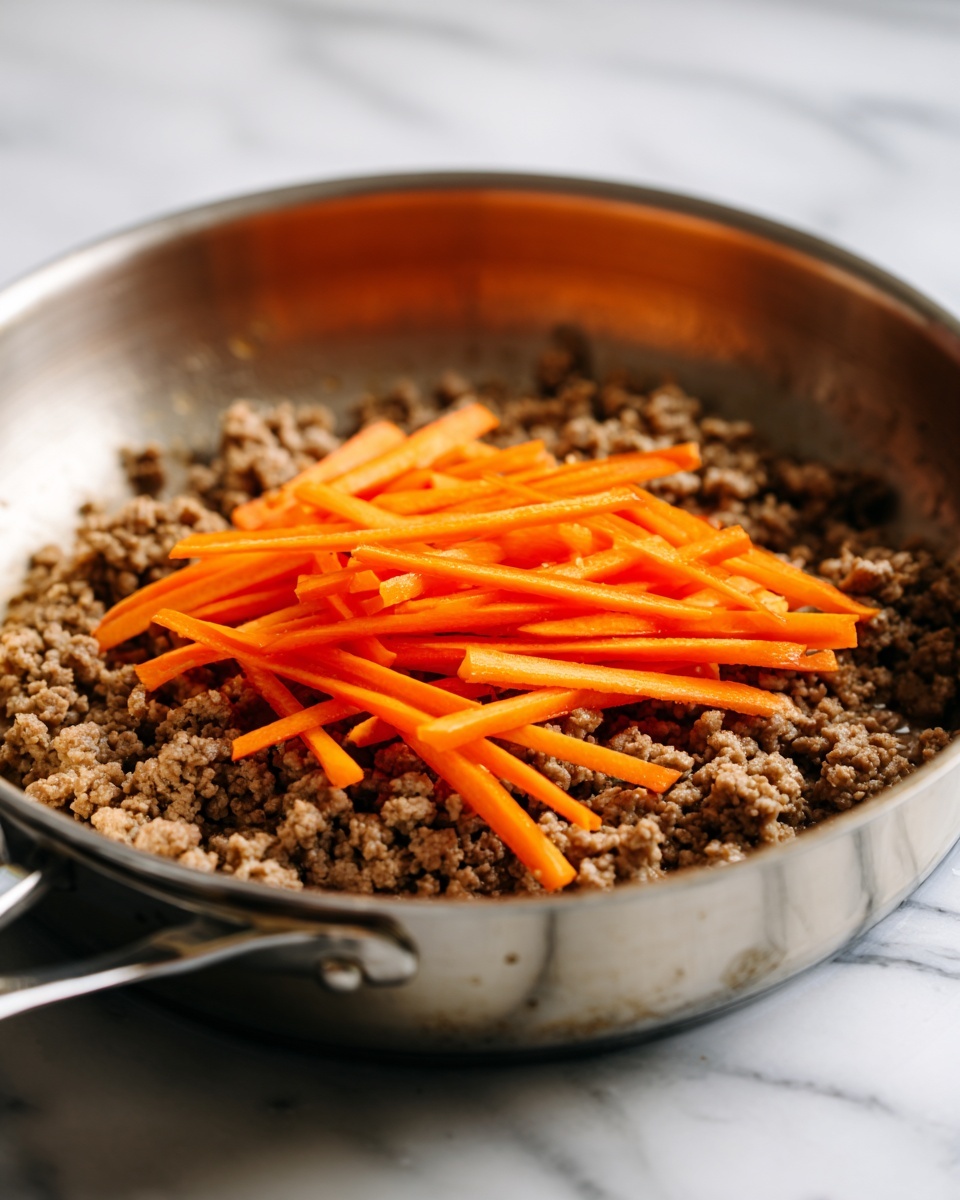A metal pan sits on a stove filled with a base layer of cooked ground meat that has a crumbly texture and brownish-gray color. On top of this meat layer, there is a pile of thin, orange carrot sticks placed roughly in the center. The pan's shiny silver surface contrasts with the food inside, and the background features a white marbled surface. photo taken with an iphone --ar 4:5 --v 7