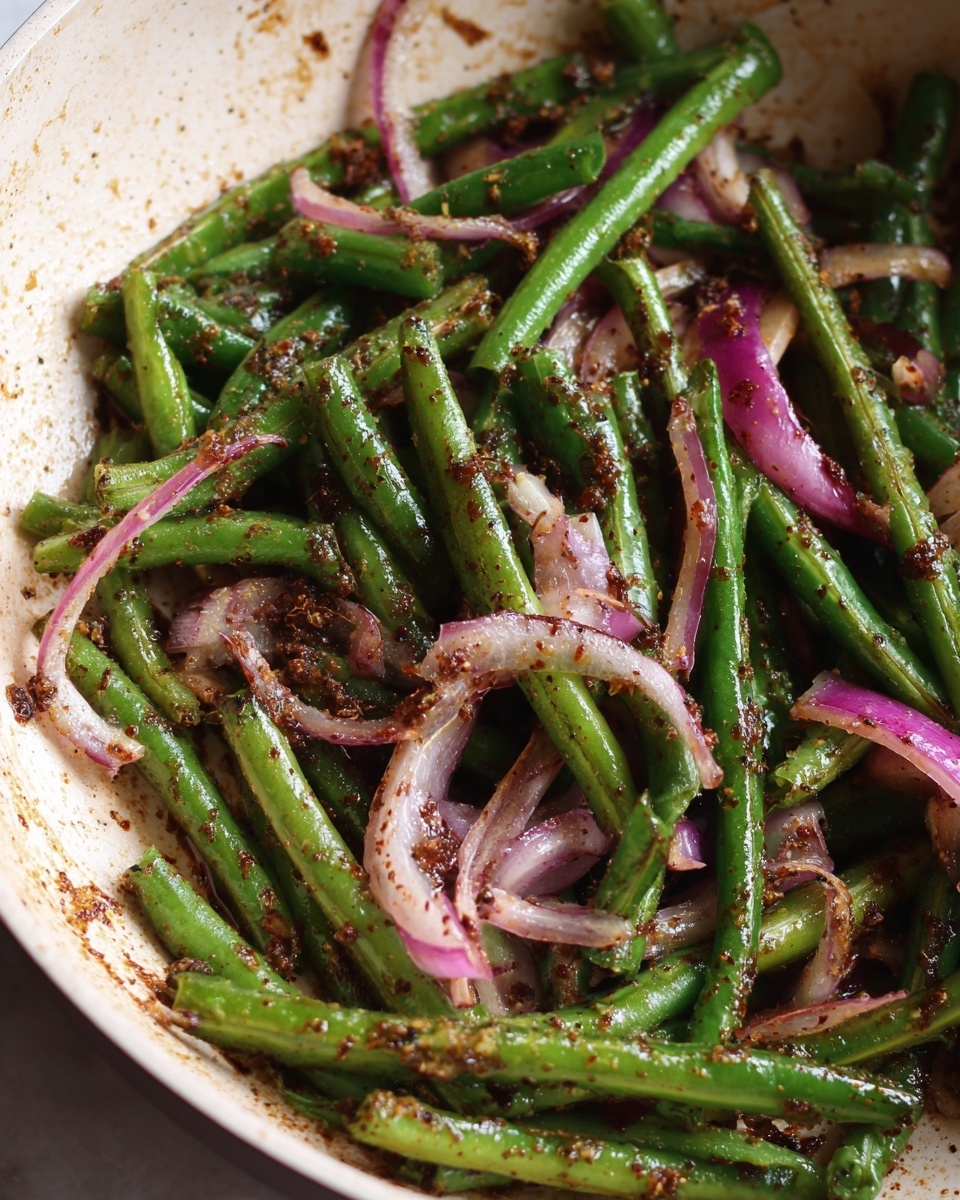 The image shows a pan with cooked green beans and slices of red onion mixed with spices. The green beans are cut into long strips, bright green in color, and slightly shiny from cooking oil. The red onions are thinly sliced and have a soft, translucent texture with a reddish-purple color. The spices, dark brown in color, are sprinkled evenly over the vegetables, creating specks and patches that add texture. The pan's inside is light-colored with some oil and spice residue. Photo taken with an iphone --ar 4:5 --v 7