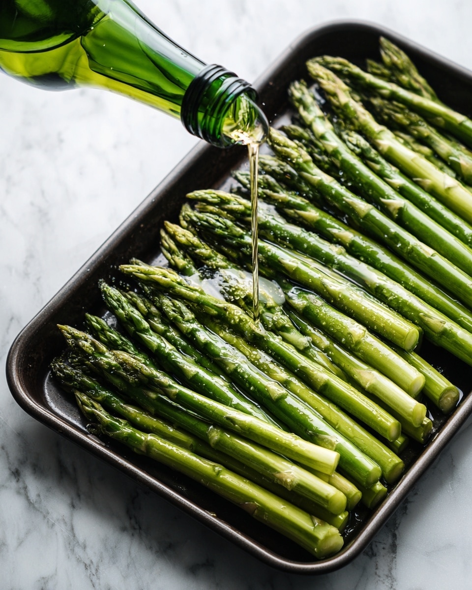 A dark metal tray holds a single layer of fresh green asparagus spears arranged neatly side by side, with their pointed tips facing the top right corner. A clear golden liquid is being poured slowly over the asparagus from a green-capped bottle held in the top left corner, glistening on the stalks. The scene is set on a white marbled surface, providing a clean and bright background. The photo is taken with an iphone --ar 4:5 --v 7