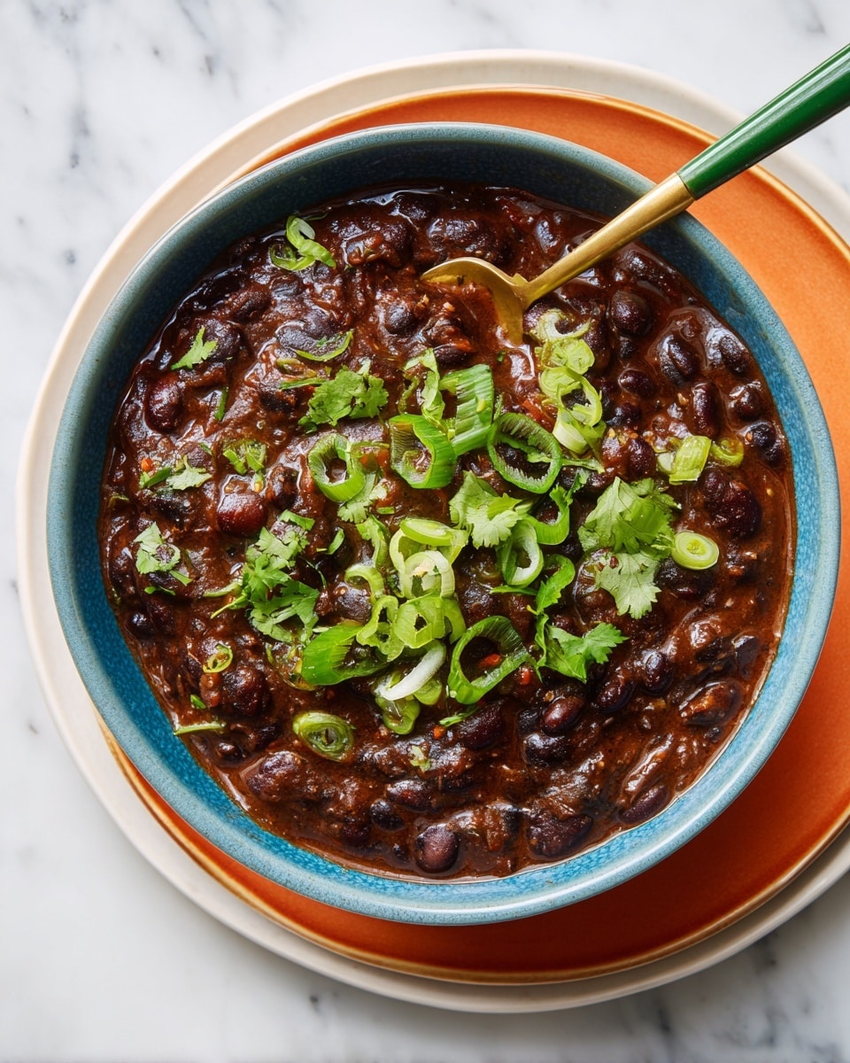 A deep blue bowl filled with thick, dark brown black bean stew, with visible whole beans and a creamy texture. The stew is topped with bright green sliced spring onions and fresh cilantro leaves scattered on top. The bowl sits on two plates, the bottom one white and the top one orange, all placed on a white marbled surface. A gold and green spoon rests inside the bowl, dipped in the stew, ready to scoop. Photo taken with an iphone --ar 4:5 --v 7