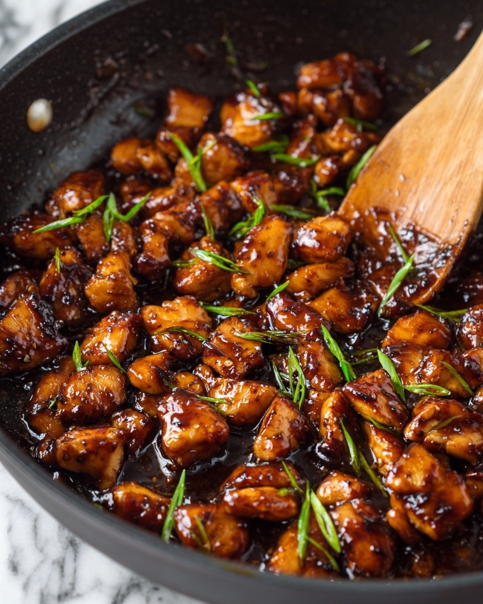 The image shows a close-up of many small pieces of cooked chicken, dark brown and shiny from sauce, with some pieces showing darker, grilled spots. The chicken pieces are mixed with small slices of green onions, adding a touch of green color. A wooden spoon with a light brown color is partly visible on the right side, stirring or resting over the chicken. All this is in a black pan, and the background is a white marbled texture. photo taken with an iphone --ar 4:5 --v 7