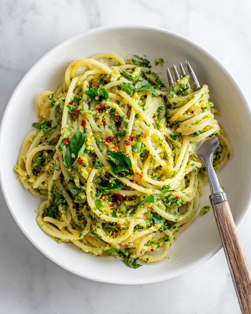 A white round plate holds a nest of thick, pale yellow spaghetti noodles mixed with a chunky, light green sauce spread throughout the pasta strands. Bright green basil and mint leaves are scattered on top, adding fresh color contrast, while small bits of red chili flakes are sprinkled lightly over the dish. Fine yellow lemon zest is also visible, giving a bright touch across the top. A fork with a silver prong and wooden handle is placed on the right side, twirling some noodles. The plate rests on a white marbled surface photo taken with an iphone --ar 4:5 --v 7