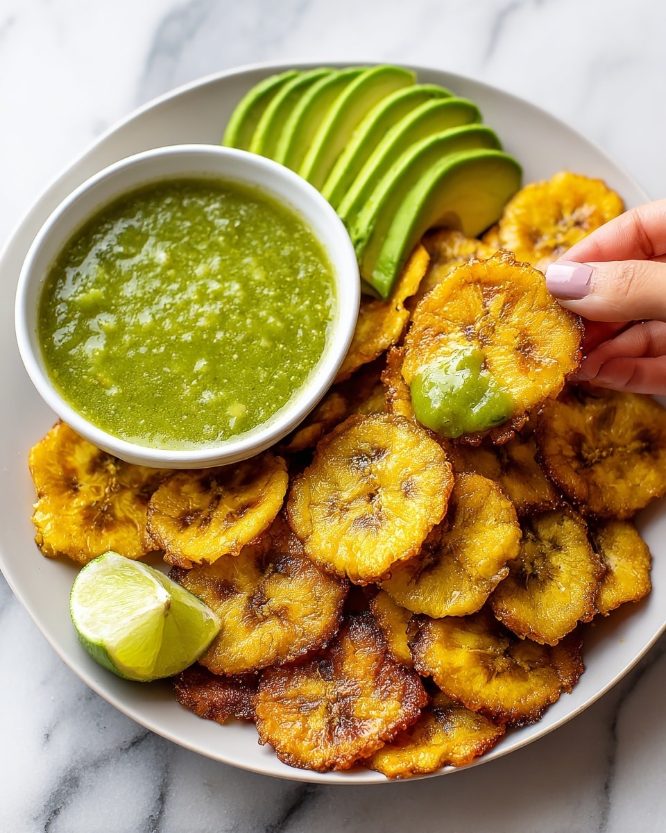 A white plate filled with crispy golden yellow fried plantain slices stacked unevenly, with a small white bowl of thick green sauce placed near the top left side of the plate next to sliced green avocado fan. A woman's hand is holding a single fried plantain slice that is dipped halfway in the green sauce. A small piece of lime wedge is visible at the bottom center of the plate. The background is a white marbled texture. photo taken with an iphone --ar 4:5 --v 7