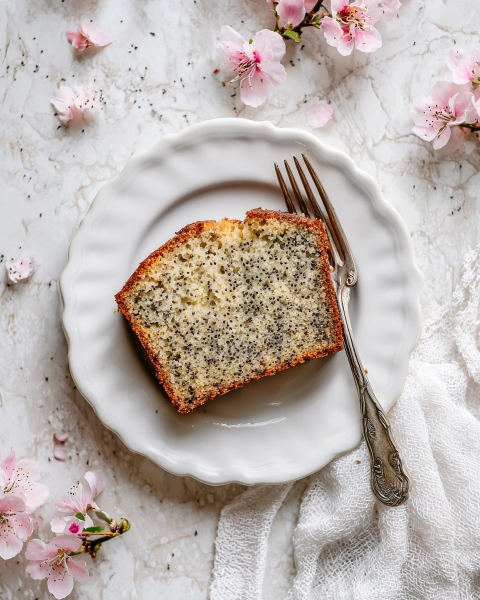 A single slice of poppy seed loaf cake with a light brown and speckled crumb sits on a white plate with scalloped edges. The cake has a thin outer crust, slightly darker brown, while the inside is soft with small black seed dots throughout. Beside the cake slice is a vintage-style silver fork lying on the plate. The plate rests on a white marbled surface scattered with delicate pink cherry blossom flowers and a white textured cloth partially visible on the right side. Photo taken with an iphone --ar 4:5 --v 7