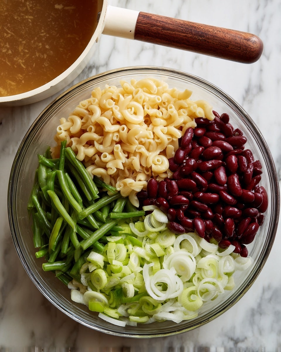 A clear glass bowl holds four separate layers of salad ingredients arranged in groups. The top left section contains shiny, red kidney beans, while the top right is filled with pale yellow cooked macaroni pasta. The bottom left part has dark green sliced green beans, and the bottom right is topped with thinly sliced pale green celery and white onion rings. Above and to the upper left of the bowl, a white pan with a wooden handle contains brownish broth or dressing. The bowl and pan are on a white marbled surface. Photo taken with an iphone --ar 4:5 --v 7