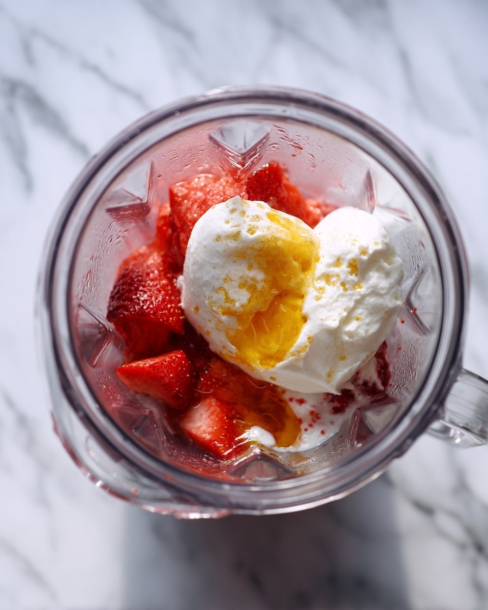 A clear blender jar is shown from above, containing two layers of ingredients: the bottom layer has frozen strawberries with a bright red, icy texture, while the top layer is a single large scoop of white creamy yogurt with a small pool of golden honey dripping into its center. The background surface is white marbled. photo taken with an iphone --ar 4:5 --v 7
