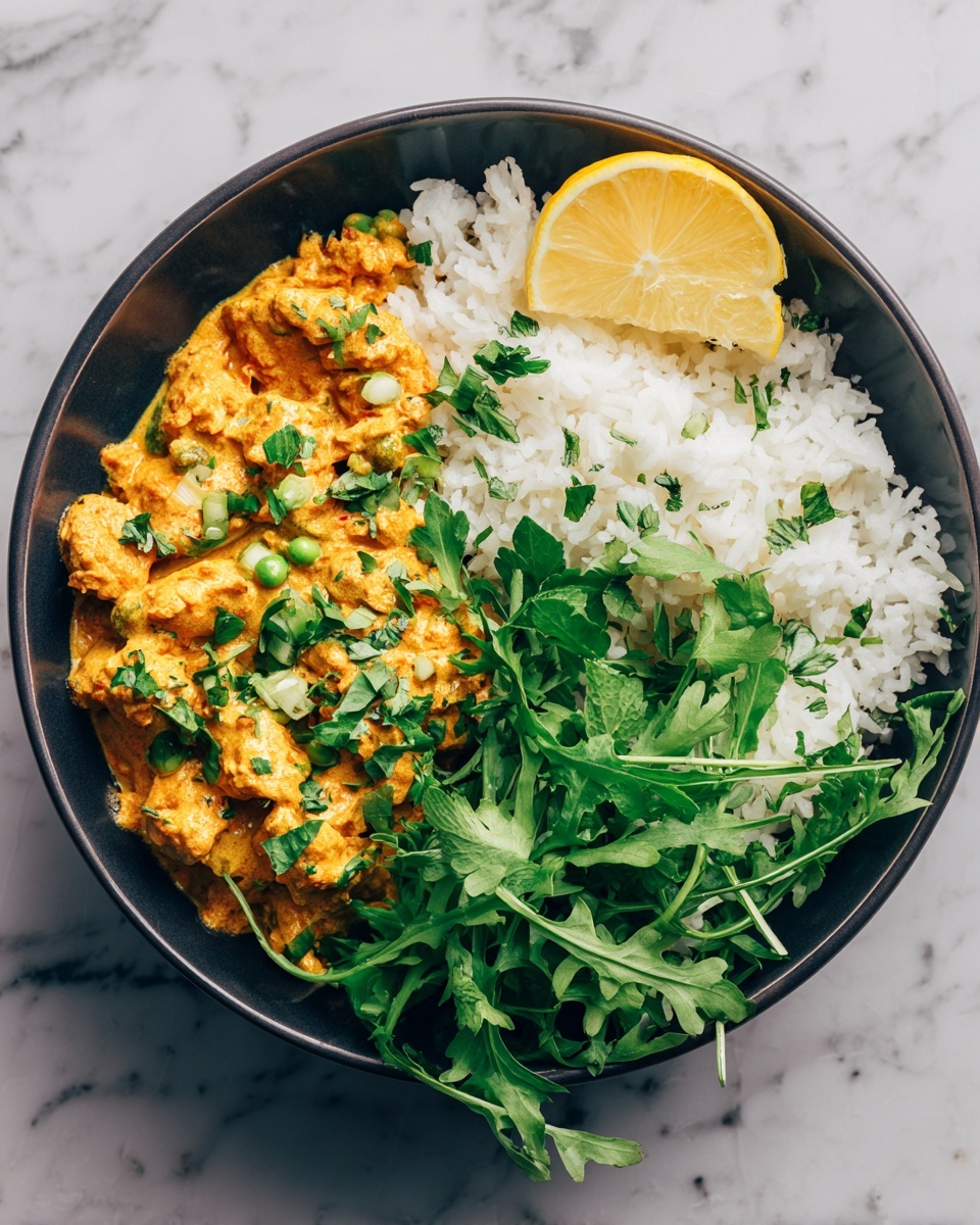 A dark bowl holds a dish with two main layers: on the left side, there is orange creamy curry mixed with green herb pieces and small vegetable chunks, topped with scattered fresh green herb leaves; on the right side, there is a fluffy white rice layer garnished with a bunch of fresh green herbs on top and a bright yellow lemon wedge resting on the edge of the bowl. The bowl sits on a white marbled textured surface, photo taken with an iphone --ar 4:5 --v 7