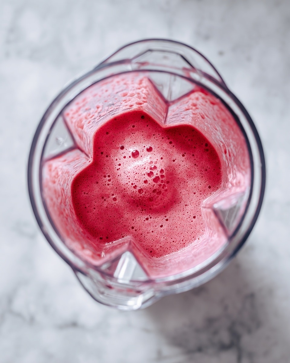 The image shows a clear blender container filled with a bright pink smoothie mix inside, showing a smooth yet slightly textured surface with light foam around the edges near the container wall. The blender container has clear geometric features and metal blades visible from the top view, with some drops on the inside surface. The scene is set on a white marbled texture surface that reflects light softly, highlighting the blender's shape and the vibrant color of the smoothie inside photo taken with an iphone --ar 4:5 --v 7