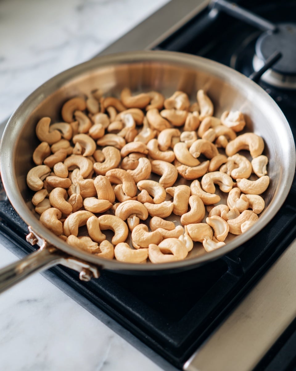 A round silver pan is shown with a single layer of light brown cashew nuts spread evenly inside. Each nut has a smooth texture with some slight color variations from beige to light tan. The pan sits on a black stove burner against a white marbled surface in the background. The cashews are whole and scattered without overlapping. The lighting is bright, creating soft shadows around the nuts, showing their curved shapes clearly. photo taken with an iphone --ar 4:5 --v 7