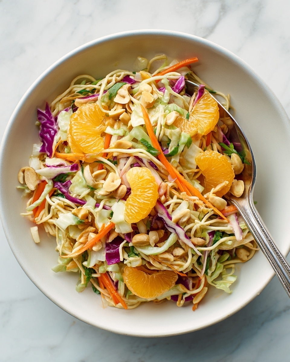 A clear white bowl holds a colorful salad with multiple layers, including orange mandarin slices, thin orange carrot strips, small bits of purple cabbage, shredded pale green cabbage, and pieces of light brown nuts scattered throughout, all mixed with small round beige crunchy noodles. The salad looks fresh and vibrant against a white marbled texture background, with a metal utensil scooping some of the salad. photo taken with an iphone --ar 4:5 --v 7