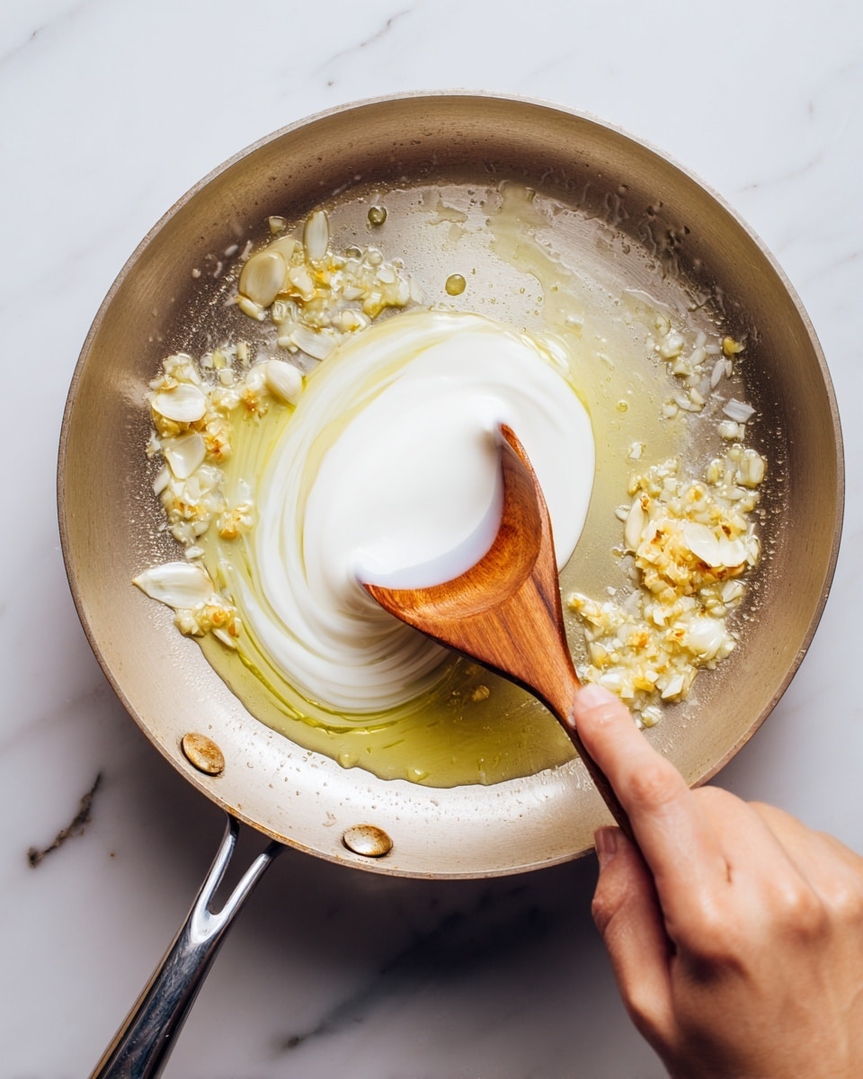 A close-up of a silver pan on a white marbled surface with melted butter and small bits of garlic or onion sizzling inside. A woman's hand is pouring thick white cream from a white measuring cup into the pan. A wooden spoon with a smooth texture rests inside the pan, partially immersed in the butter and cream mixture, stirring gently. Photo taken with an iphone --ar 4:5 --v 7