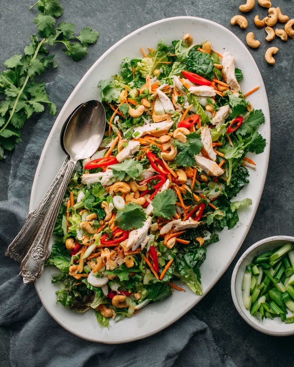 A white oval plate holds a colorful salad with several layers. The base layer is a mix of green leafy vegetables including chopped lettuce and some dark green leaves. On top, there are thin orange carrot strips and long red pepper slices scattered throughout. Pieces of light tan cooked chicken are spread over the salad, along with whole light brown cashew nuts. Fresh green onion slices and cilantro leaves add bright green pops across the dish. A shiny silver spoon and fork rest on the left side of the plate. The plate sits on a dark gray surface with a few green onion slices and a small white bowl filled with green onions on the bottom right. photo taken with an iphone --ar 4:5 --v 7