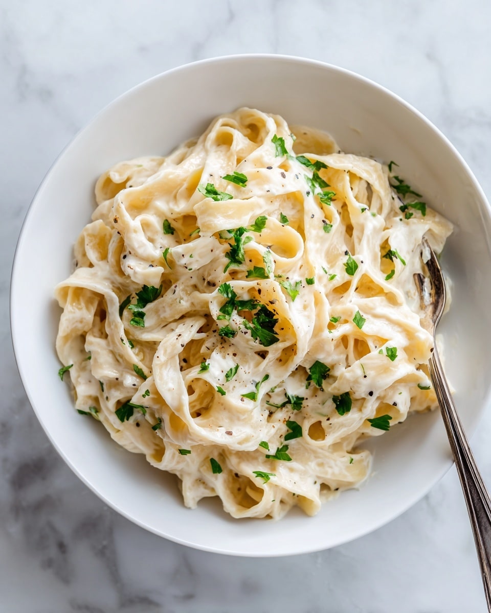 A white bowl holds creamy fettuccine pasta coated evenly in a smooth, light ivory sauce. The pasta noodles are thick and flat, twirled together to create layers of soft texture. Small flecks of green parsley are scattered on top and around the pasta, adding a fresh touch of color. A silver fork is partly inserted into the pasta in the bowl. The bowl sits on a white marbled surface. photo taken with an iphone --ar 4:5 --v 7