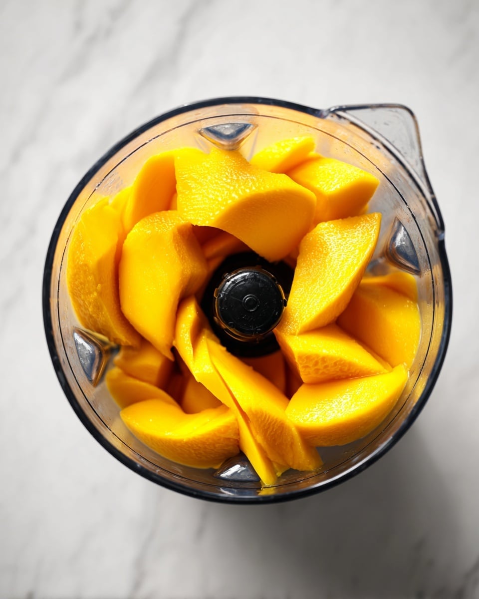 A clear blender container filled with bright yellow mango slices arranged around the black central blade, showing smooth and large pieces piled up inside. The background is a white marbled surface. photo taken with an iphone --ar 4:5 --v 7
