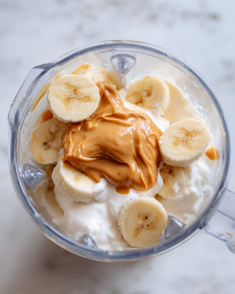 The image shows a close-up of a clear glass blender jar with visible metal blades at the bottom, filled with a mixture of sliced banana pieces, a dollop of light brown peanut butter, and a soft white creamy substance, all resting on a white marbled surface. The banana slices are pale yellow, round, and placed on the top and around the peanut butter, while the peanut butter sits in the center, smooth and creamy, slightly oozing onto the white cream beneath it, creating a soft texture contrast. The scene is bright and clear, capturing the freshness and creamy textures of the ingredients. Photo taken with an iphone --ar 4:5 --v 7