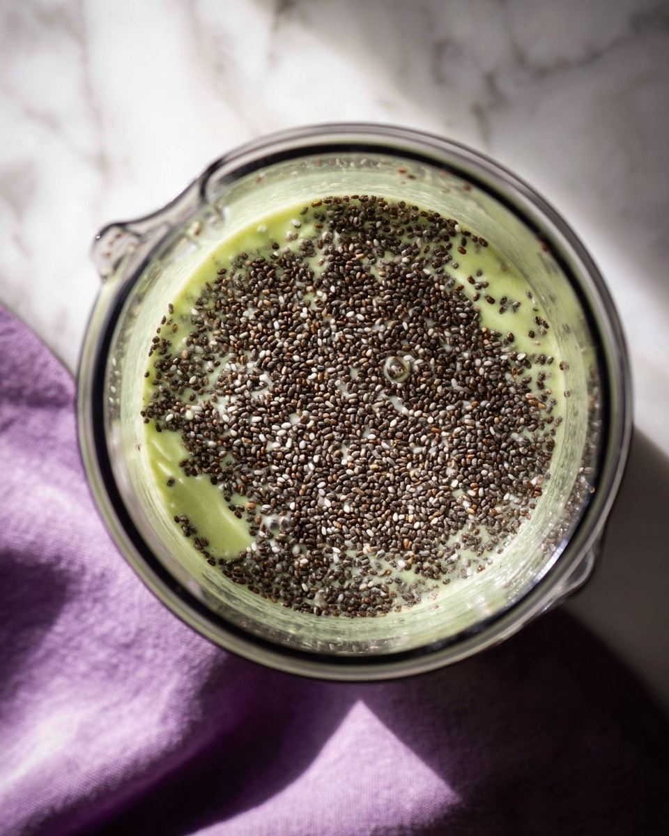 The image shows a clear blender jar viewed from above with many small black chia seeds floating on top of a thin light green liquid layer. The chia seeds cover the entire visible surface inside the blender, creating a textured, dense black layer with tiny hints of white from some seeds. The blender is sitting on a white marbled surface with part of a purple cloth visible at the bottom edge. The natural light casts soft shadows inside the blender, highlighting the contrast between the dark seeds and pale liquid photo taken with an iphone --ar 4:5 --v 7