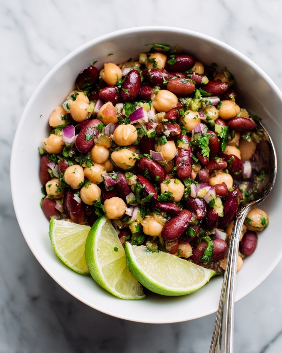 A white bowl holds a colorful bean salad made of three types of beans: red kidney beans, light brown beans, and chickpeas, all mixed evenly with chopped green herbs and small pieces of red onion, adding green and purple colors. There are two lime wedges placed at the front inside the bowl. A shiny silver spoon rests on the right side, partially inside the bowl. The background is a white marbled texture. photo taken with an iphone --ar 4:5 --v 7
